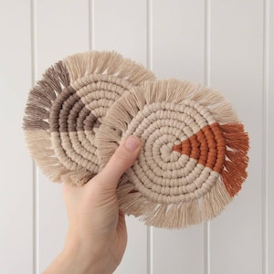 May include: Two macrame coasters are held in a hand. The coasters are circular with a spiral design and fringed edges. One has a brown and beige colour block, the other a rust and beige colour block. The background is a white wall.