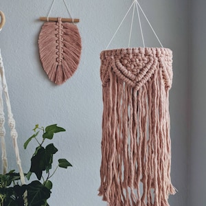 Puede incluir: Un móvil colgante de macramé rosa con un anillo de madera en la parte superior y una franja de borlas. El móvil está colgado de un cordón blanco. Hay una pequeña pared de macramé rosa en el fondo.