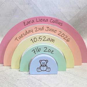 May include: A multi-coloured wooden rainbow stacker featuring five arched segments in pink, peach, yellow, mint green, and light blue. The segments are printed with the name Zara Liena Collins, the date Tuesday 2nd June 2026, 10.52am, 7lb 2oz, and a teddy bear.
