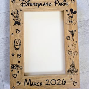May include: Wooden picture frame with the words "Disneyland Paris" and "March 2026" etched in black. The frame is decorated with cartoon characters and icons, including Mickey and Minnie Mouse, and a castle.