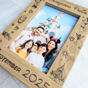 May include: Wooden photo frame with a family portrait in front of a castle. The frame is engraved with Disney-themed illustrations and the text "Disneyland Paris" and "September 2025". The frame has a light brown color.