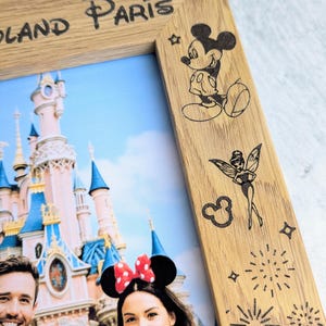 May include: Wooden photo frame with the words "Disneyland Paris" etched at the top. The frame features etched images of Mickey Mouse, Tinkerbell, and fireworks. The photo inside shows a couple in front of a castle.