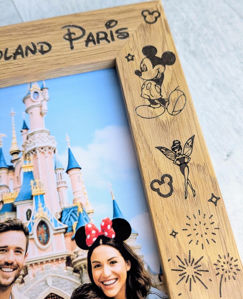 May include: Wooden photo frame with the words "Disneyland Paris" etched at the top. The frame features etched images of Mickey Mouse, Tinkerbell, and fireworks. The photo inside shows a couple in front of a castle.
