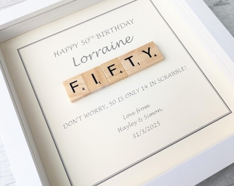 May include: A white framed picture with a cream background. The picture says "Happy 50th Birthday Lorraine" and features the word "FIFTY" spelled out with Scrabble tiles. The text below the tiles says "Don't worry, 50 is only 14 in Scrabble!" and is signed "Love from Hayley & Simon. 31/3/2025."