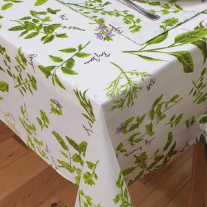 Puede incluir: Mantel blanco con estampado de hierbas verdes y marrones. El mantel presenta los nombres de diferentes hierbas, incluyendo albahaca, orégano, menta y tomillo.
