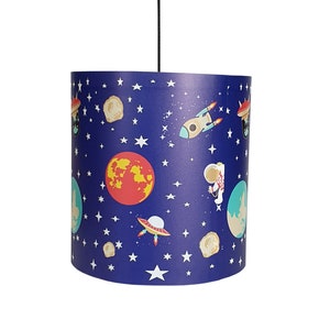 Może przedstawiać: Abażur lampy w kolorze niebieskim i białym z motywem kosmicznym, przedstawiający rakietę, planety, gwiazdy i astronautę.
