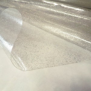 May include: Clear plastic table cover with a subtle sparkle finish. The cover is folded in half, showing the texture of the material.