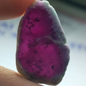 May include: A rough gemstone with a vivid magenta core and a pale green and grey outer edge. The stone is held between fingers, displaying its natural, irregular shape and unique colour variations. The gem's surface has dark inclusions.
