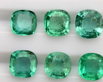 May include: Six cushion-cut emerald gemstones. The gems are a vibrant green color, with visible facets and a polished surface. The stones are arranged in two rows of three, showcasing their clarity and brilliance.