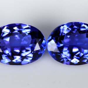 May include: Two oval-cut, faceted gemstones with a deep blue hue. The gems have a brilliant, reflective surface, showcasing internal facets that catch and reflect light. The stones are set against a plain white background.