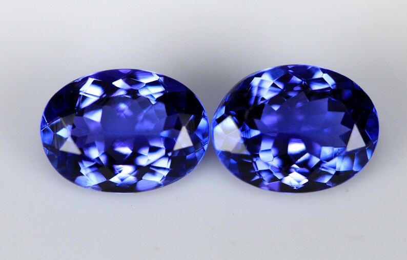 May include: Two oval-cut, faceted gemstones with a deep blue hue. The gems have a brilliant, reflective surface, showcasing internal facets that catch and reflect light. The stones are set against a plain white background.