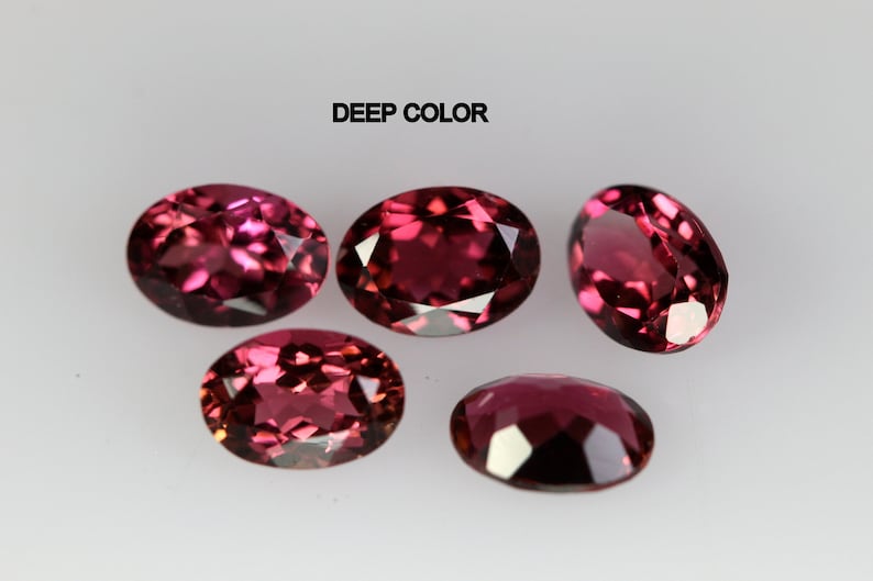 May include: Five oval-shaped gemstones with a deep red color. The gemstones are arranged in a semi-circle on a white surface. The text "DEEP COLOR" is printed above the gemstones.