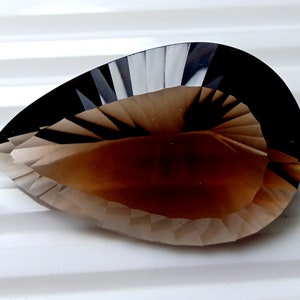 May include: A faceted, pear-shaped gemstone with a gradient of colors, transitioning from a dark, almost black, border to a light brown center. The gem's surface is cut with precise angles, creating a play of light and shadow.