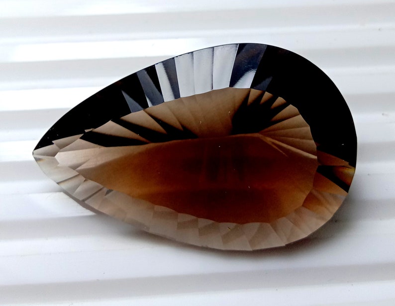 May include: A faceted, pear-shaped gemstone with a gradient of colors, transitioning from a dark, almost black, border to a light brown center. The gem's surface is cut with precise angles, creating a play of light and shadow.