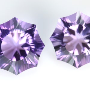 May include: Two faceted, starburst-shaped gemstones in shades of lavender and purple. The gems have a central circular facet surrounded by radiating, triangular facets. The background is a plain white.