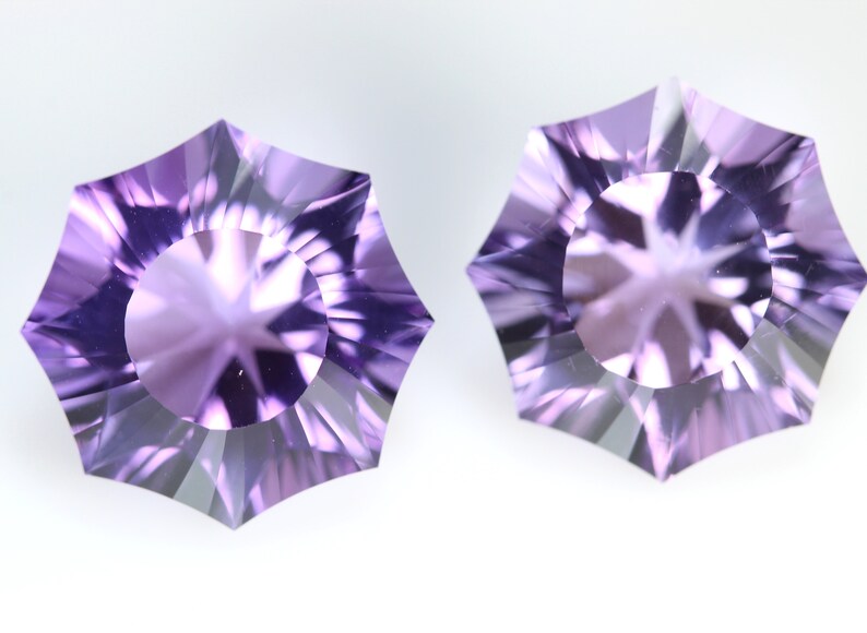 May include: Two faceted, starburst-shaped gemstones in shades of lavender and purple. The gems have a central circular facet surrounded by radiating, triangular facets. The background is a plain white.