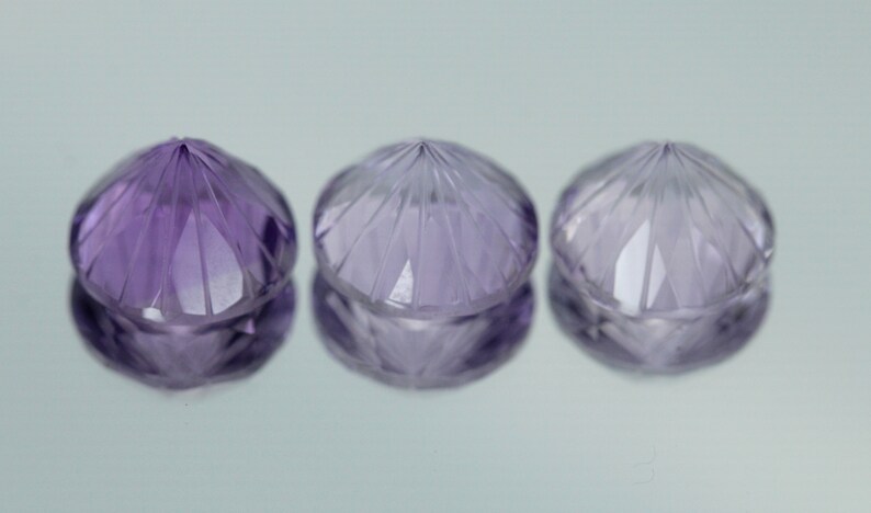 May include: Three faceted amethyst gemstones, each with a pointed top and a round base. The gemstones are a light purple color and have a shiny, polished finish.