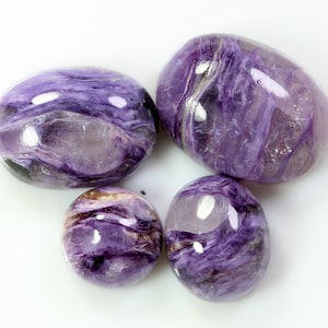 Puede incluir: Cuatro piedras de charoíta pulidas de diferentes tamaños. Las piedras muestran un patrón arremolinado de tonos morados oscuros y lavanda, con algunas áreas translúcidas. Las superficies pulidas reflejan la luz, resaltando los patrones y colores naturales.