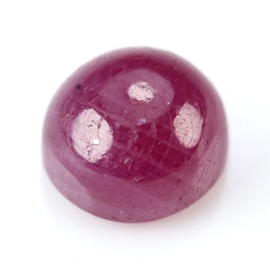 May include: A polished, cabochon-cut ruby gemstone. The deep red gem has a smooth, rounded surface with subtle internal inclusions and a glossy sheen. The ruby is set against a bright white background.