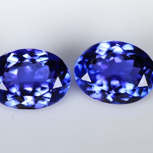 May include: Two oval-cut, faceted gemstones with a deep blue color. The gems have a brilliant, reflective surface, showcasing a complex pattern of internal facets. The stones are set against a plain white background.