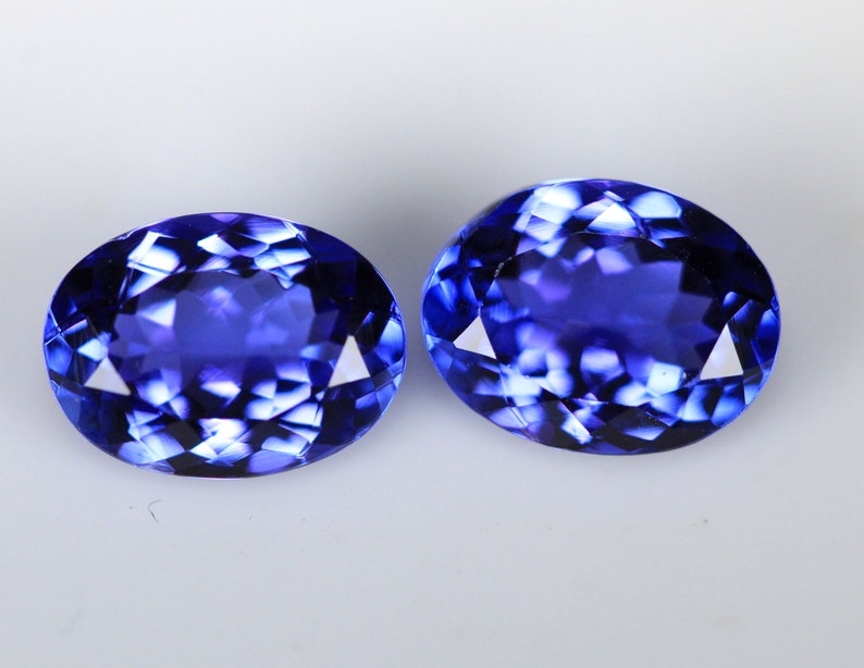 May include: Two oval-cut, faceted gemstones with a deep blue color. The gems have a brilliant, reflective surface, showcasing a complex pattern of internal facets. The stones are set against a plain white background.
