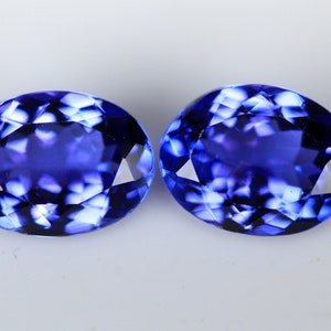 May include: Two oval-cut, faceted gemstones with a deep blue color. The gems have a high degree of clarity and brilliance, reflecting light to create a sparkling effect. The stones are set against a plain white background.