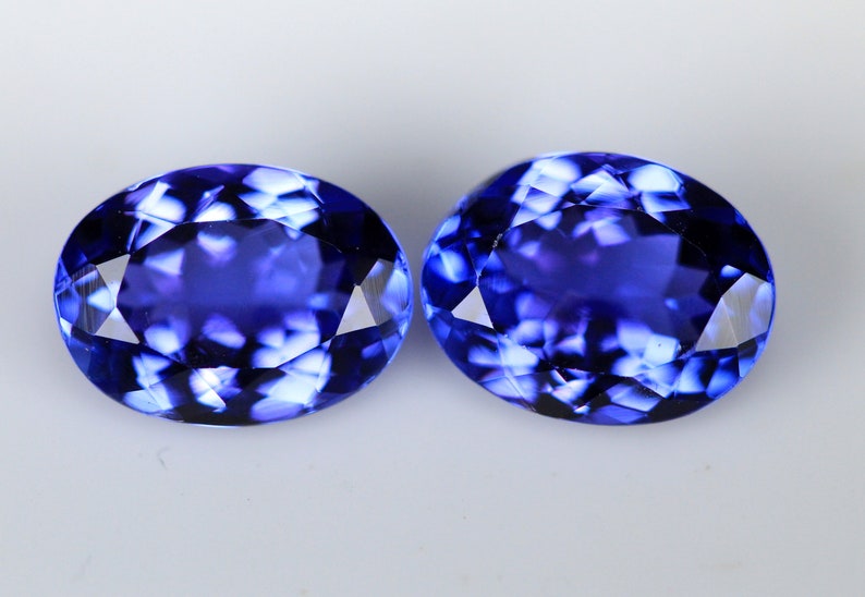 May include: Two oval-cut, faceted gemstones with a deep blue color. The gems have a high degree of clarity and brilliance, reflecting light to create a sparkling effect. The stones are set against a plain white background.