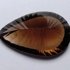 May include: A faceted, teardrop-shaped gemstone with a gradient of brown hues. The gem has a polished surface with precise cuts that reflect light, creating a sparkling effect. The stone's shape is elongated, with a pointed tip.