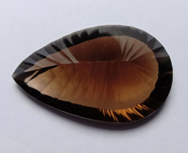 May include: A faceted, teardrop-shaped gemstone with a gradient of brown hues. The gem has a polished surface with precise cuts that reflect light, creating a sparkling effect. The stone's shape is elongated, with a pointed tip.