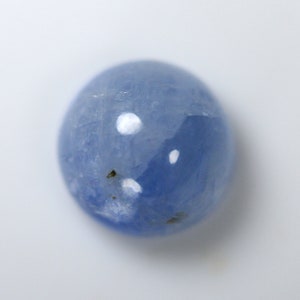 Puede incluir: Una gema ovalada lisa de color azul claro. La gema tiene una superficie pulida con algunas pequeñas inclusiones oscuras y reflejos blancos brillantes. La piedra está sobre un fondo blanco liso.