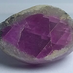 Op de afbeelding: Een gefacetteerde, ovaalvormige edelsteen met een diep magenta interieur en een bleke, grijswitte buitenlaag. Het oppervlak van de steen vertoont een geometrisch patroon dat licht reflecteert. De edelsteen is waarschijnlijk een gepolijst mineraal.