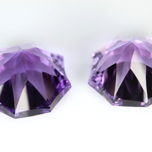 May include: Two faceted, star-shaped gemstones in shades of purple. The gems have multiple facets that reflect light, creating a sparkling effect. The gemstones are set against a white background.