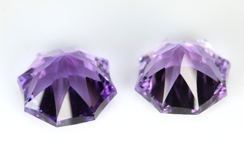 May include: Two faceted, star-shaped gemstones in shades of purple. The gems have multiple facets that reflect light, creating a sparkling effect. The gemstones are set against a white background.