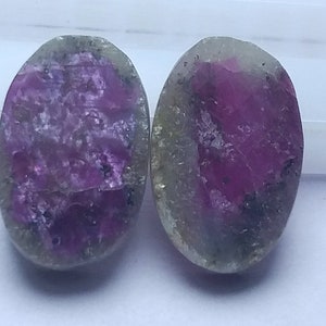 Può includere: Due gemme ovali con un vivace centro rosa e uno strato esterno traslucido, verde-grigio. Le pietre lucide mostrano motivi e inclusioni naturali. Le gemme sono su uno sfondo bianco.