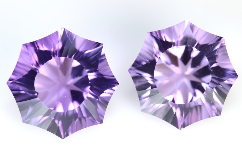 May include: Two faceted, star-shaped gemstones with a light purple hue. The gems have a central circular facet surrounded by a series of triangular facets, creating a starburst effect. The background is plain white.