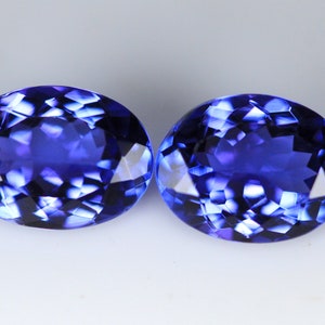 May include: Two oval-cut, deep blue gemstones with multifaceted surfaces. The gems are displayed against a white background, showcasing their brilliant, reflective qualities. The stones are similar in size and shape, suggesting they are a matched pair.