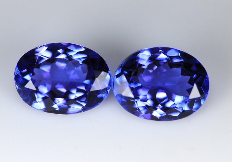 May include: Two oval-cut, deep blue gemstones with multifaceted surfaces. The gems are displayed against a white background, showcasing their brilliant, reflective qualities. The stones are similar in size and shape, suggesting they are a matched pair.