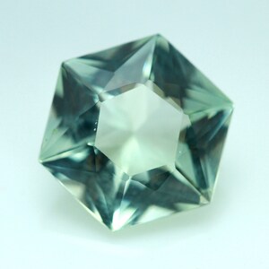 May include: A faceted gemstone with a complex, multi-sided shape. The gem has a pale green hue with internal facets that reflect light. The overall form is geometric, with sharp angles and a central, lighter-colored core.