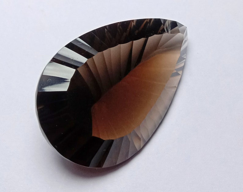 May include: A faceted, teardrop-shaped gemstone with a gradient of brown and smoky gray hues. The gem's surface is cut with precise angles, creating a play of light and shadow. The overall effect is a polished, elegant appearance.