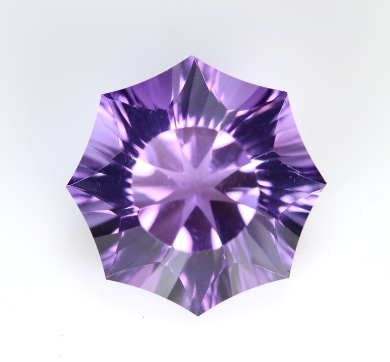 May include: A faceted, starburst-shaped gemstone in shades of purple. The gem has a symmetrical design with pointed edges radiating from a central point. The facets create a play of light and shadow, enhancing its brilliance.