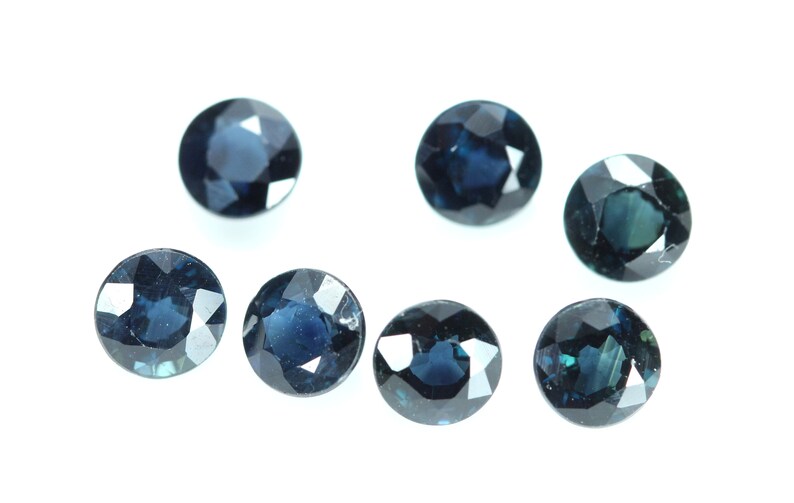 May include: Six round, dark blue gemstones with faceted edges. The gemstones are arranged in a semi-circle on a white background.