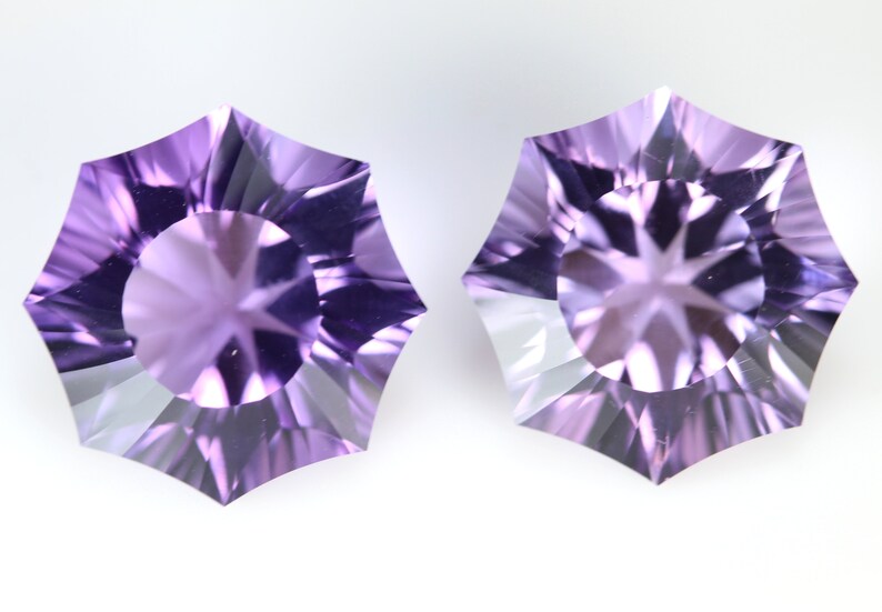 May include: Two faceted, star-shaped gemstones with a light purple hue. The gems have a central circular facet surrounded by radiating facets, creating a starburst effect. The gems are displayed against a white background.
