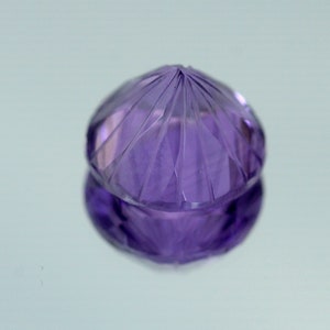 May include: A purple amethyst gemstone with a faceted cut. The gemstone is round and has a pointed top.