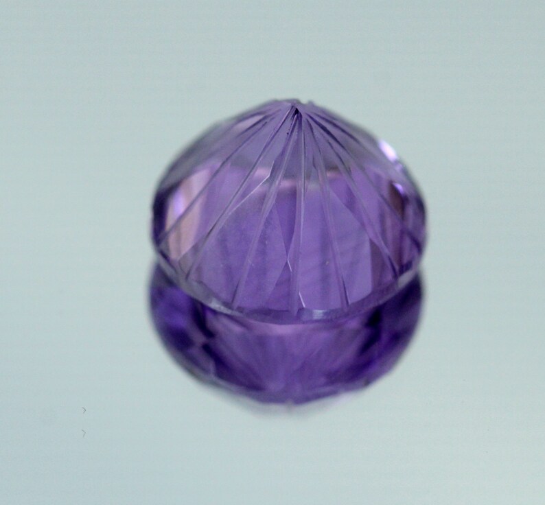 May include: A purple amethyst gemstone with a faceted cut. The gemstone is round and has a pointed top.
