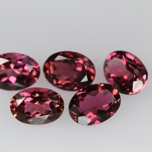 May include: Five oval-shaped gemstones, each with a deep pink hue and a faceted cut. The gemstones are arranged in a cluster on a white surface.