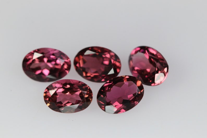 May include: Five oval-shaped gemstones, each with a deep pink hue and a faceted cut. The gemstones are arranged in a cluster on a white surface.