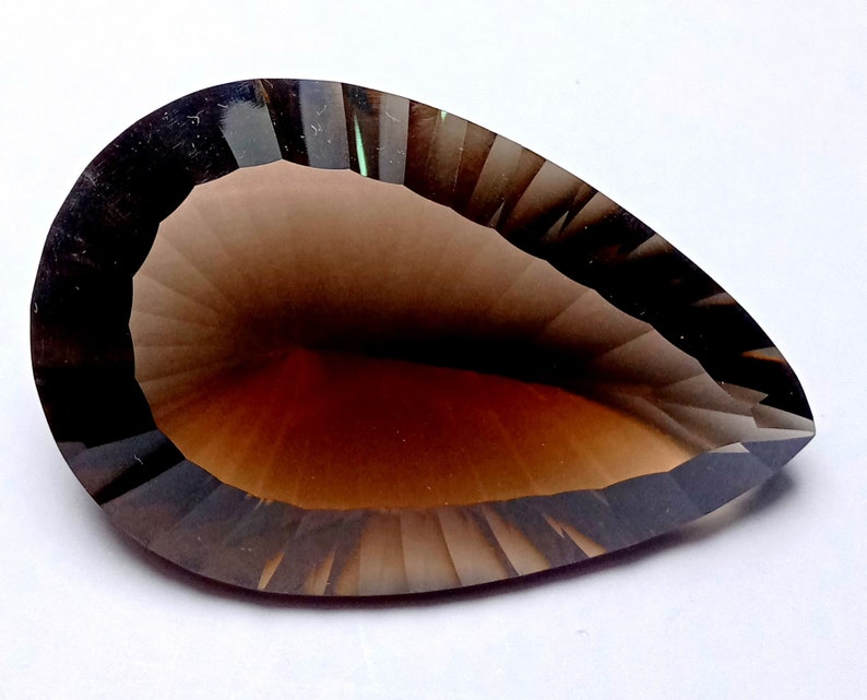 May include: A faceted, pear-shaped gemstone with a gradient of brown hues. The gem has a dark, almost black, border that transitions to a lighter brown center. The cut of the stone creates a play of light and shadow.