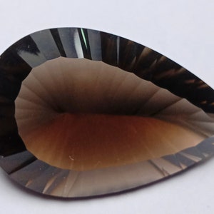 May include: A faceted, pear-shaped gemstone with a smoky brown hue. The gem has a polished surface with multiple facets, creating a play of light and shadow. The overall shape is elongated, with a rounded base and a pointed tip. The gem is set against a white background.