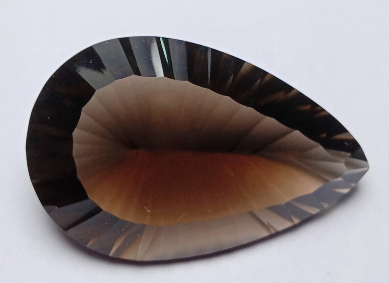 May include: A faceted, pear-shaped gemstone with a smoky brown hue. The gem has a polished surface with multiple facets, creating a play of light and shadow. The overall shape is elongated, with a rounded base and a pointed tip. The gem is set against a white background.