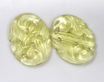 May include: Two pale yellow, oval-shaped buttons with intricate carved floral and swirl designs. Each button has six small holes for sewing. The buttons are made of a translucent material.